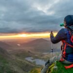 man sitting on cliffhanger looking at mountain under gloomy clouds